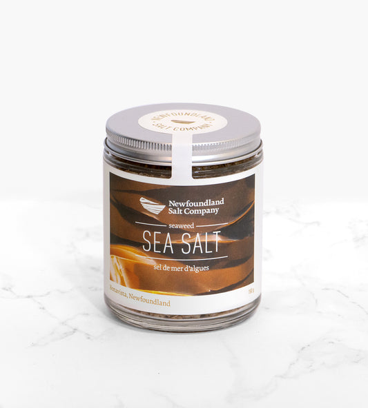 A cylindrical glass jar of a seaweed sea salt, with a silver lid. The jar is atop a white marble surface. The label on the jar depicts several strands of brown kelp.