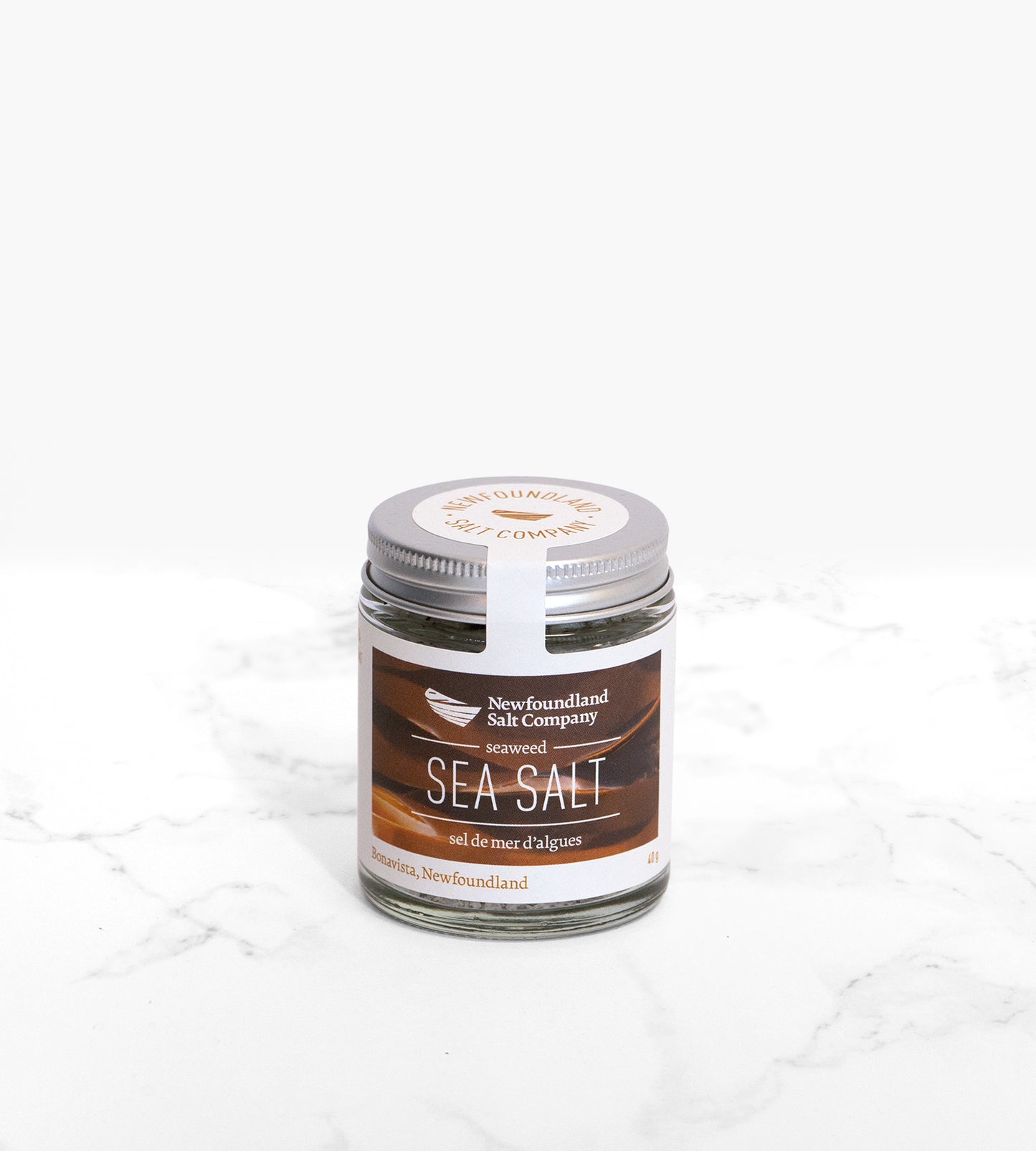 A jar of seaweed flavoured sea salt with a silver lid, on a white marble counter. The label of the jar features ribbons of brown kelp.