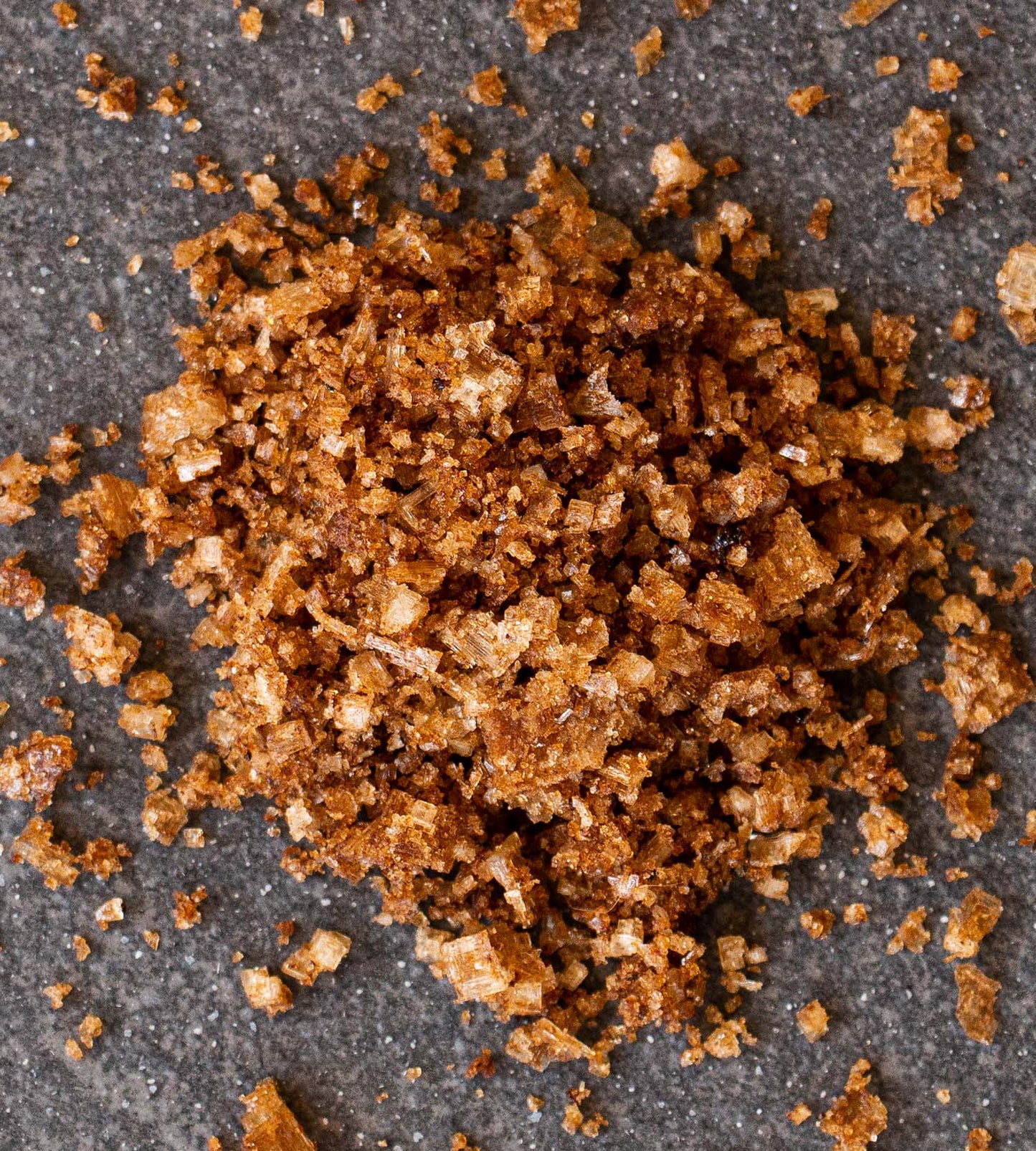 Closeup of coffee-flavoured sea salt on a dark grey tile. The flakes of salt a rich medium brown colour.