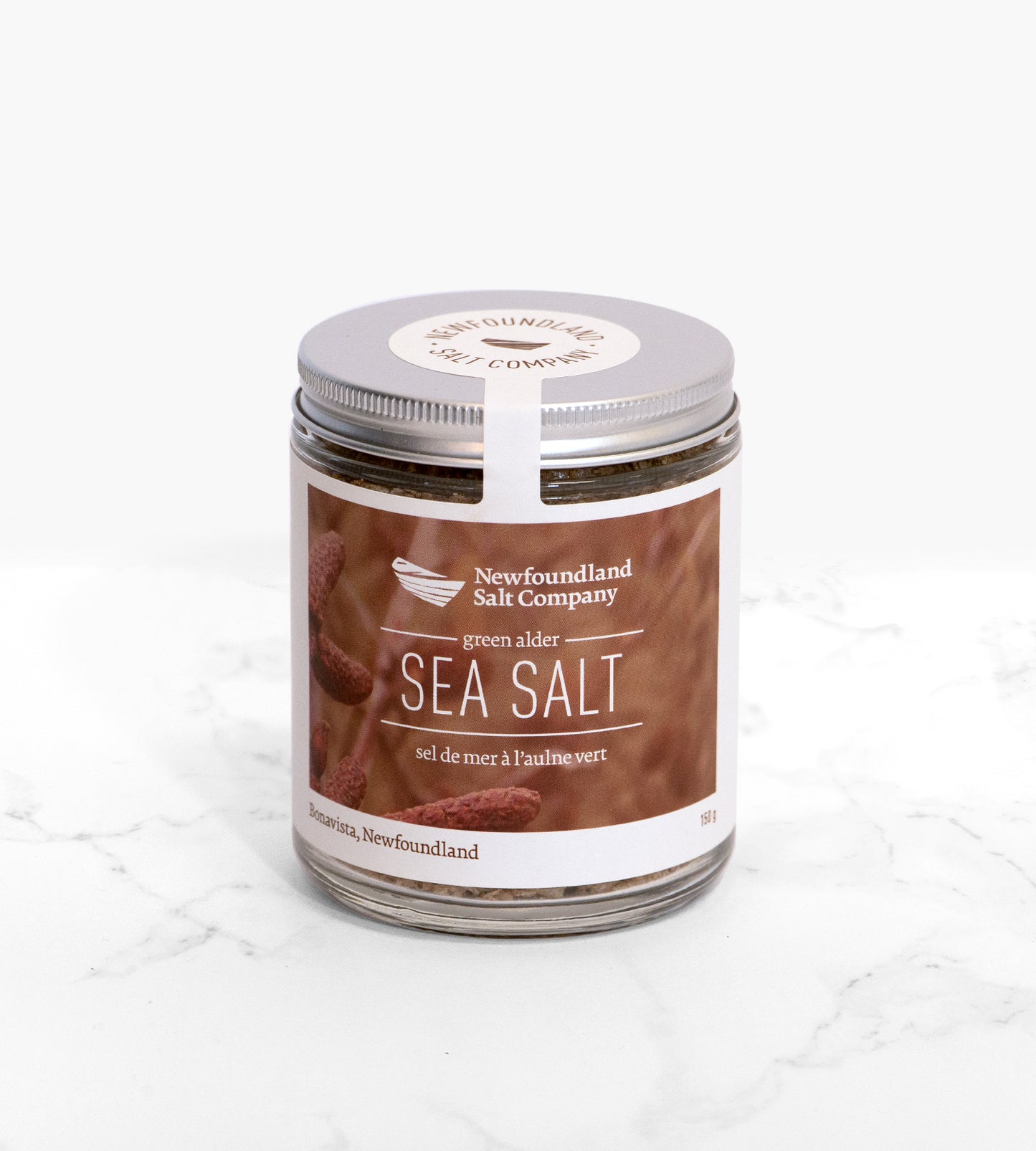 A cylindrical glass jar of sea salt flavoured with green alder, on a white marble tile. The label features a textural brown photo of alder cones.