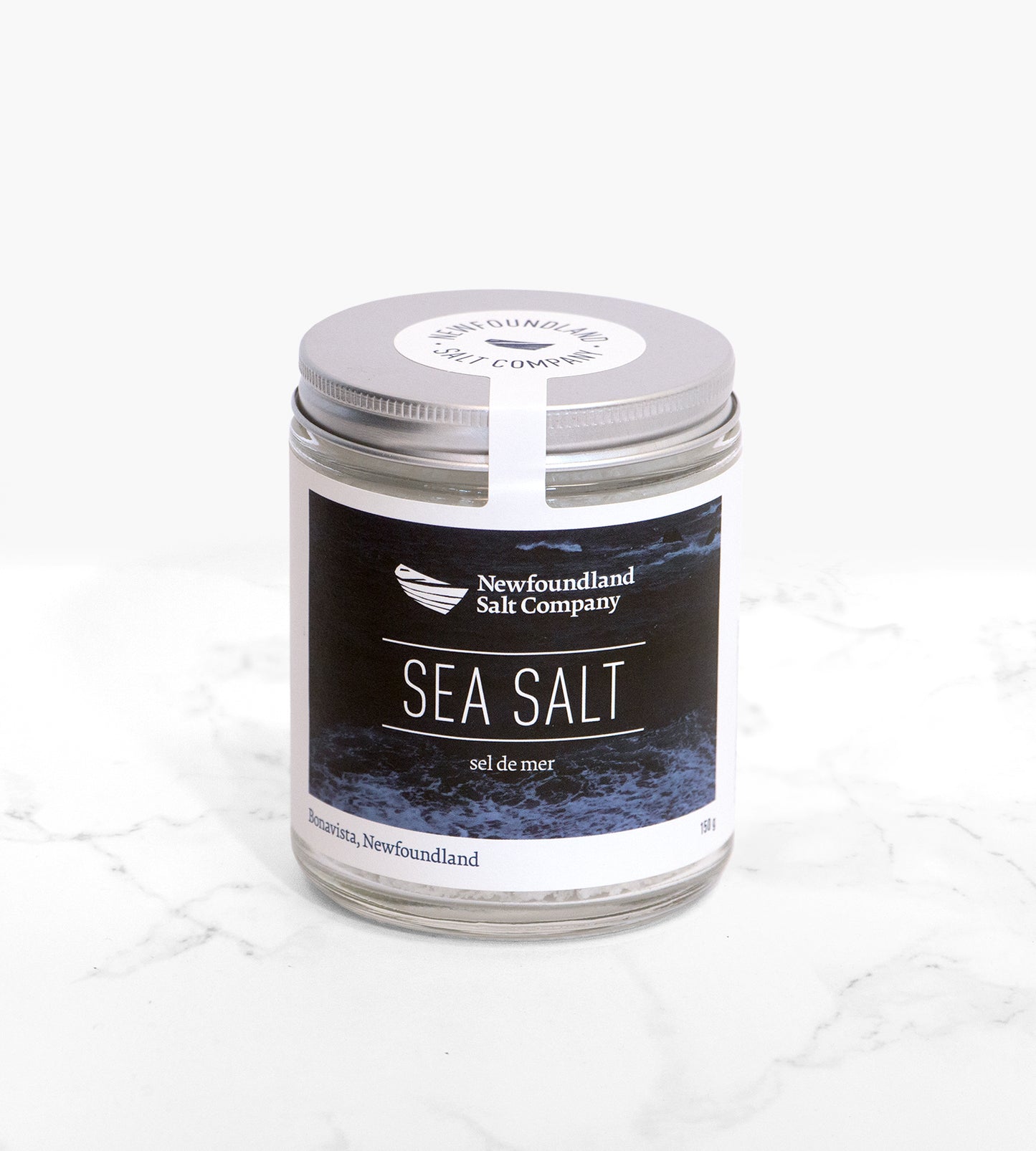 A cylindrical glass jar of sea salt flakes, with a silver lid, sits on a white marble tile. The label on the jar features a dark blue ocean wave photo.