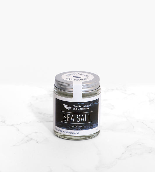 A cylindrical glass jar of sea salt, with a silver lid, sits on a white marble tile. The label on the jar features a dark blue ocean wave photo.