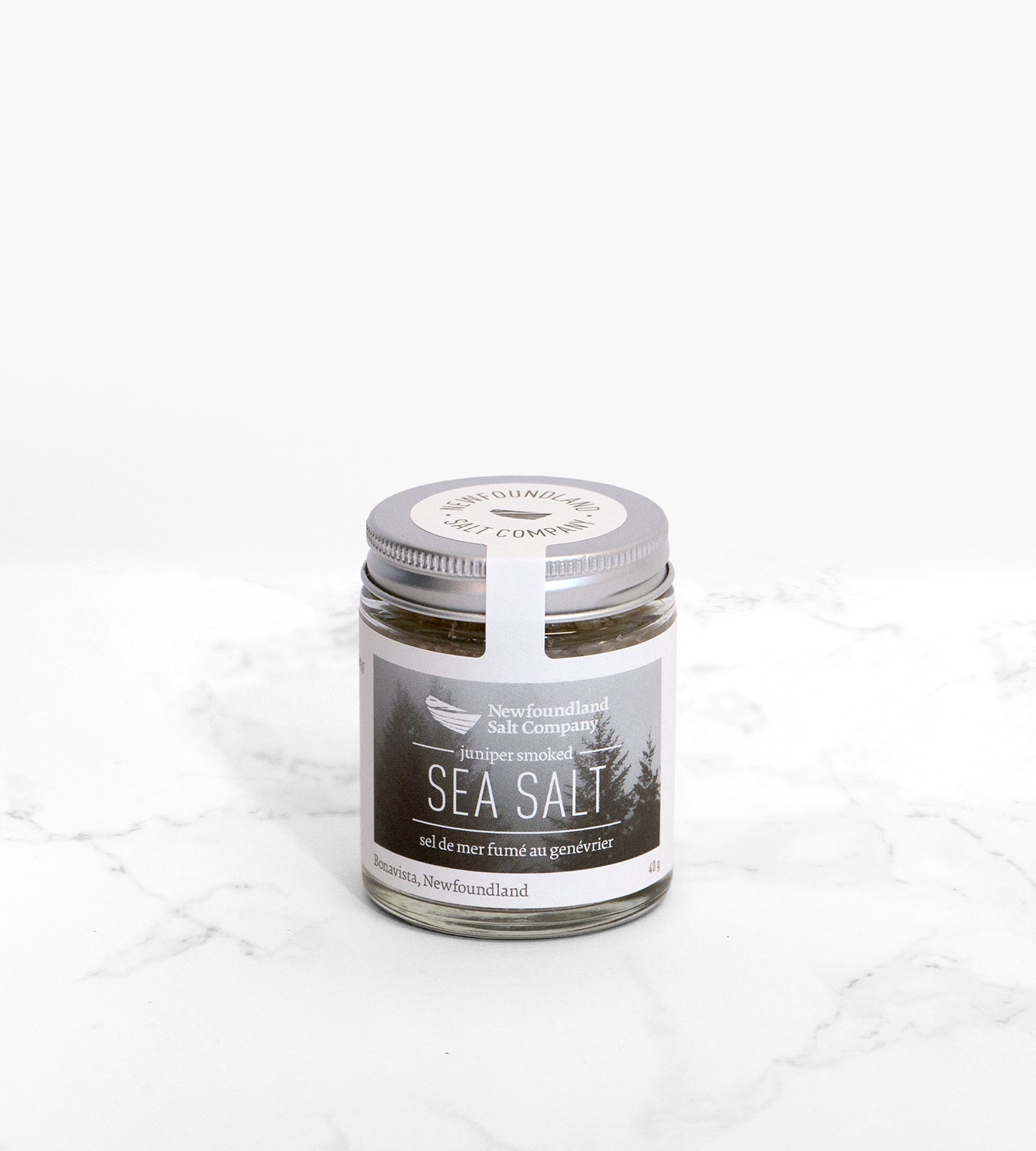 A glass jar of juniper-smoked sea salt on a white marble surface. The label on the jar depicts a foggy conifer forest.