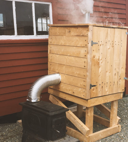 Photo of a wooden box built of planks of light-coloured wood, sitting outside a building with maroon siding. The box is connected by an aluminum hose to a woodstove, and smoke is rising from the top of the box.