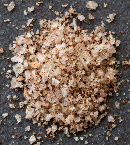 Closeup of sea salt flakes with flecks of crushed alder. The small pile of salt is on a dark grey slate tile.
