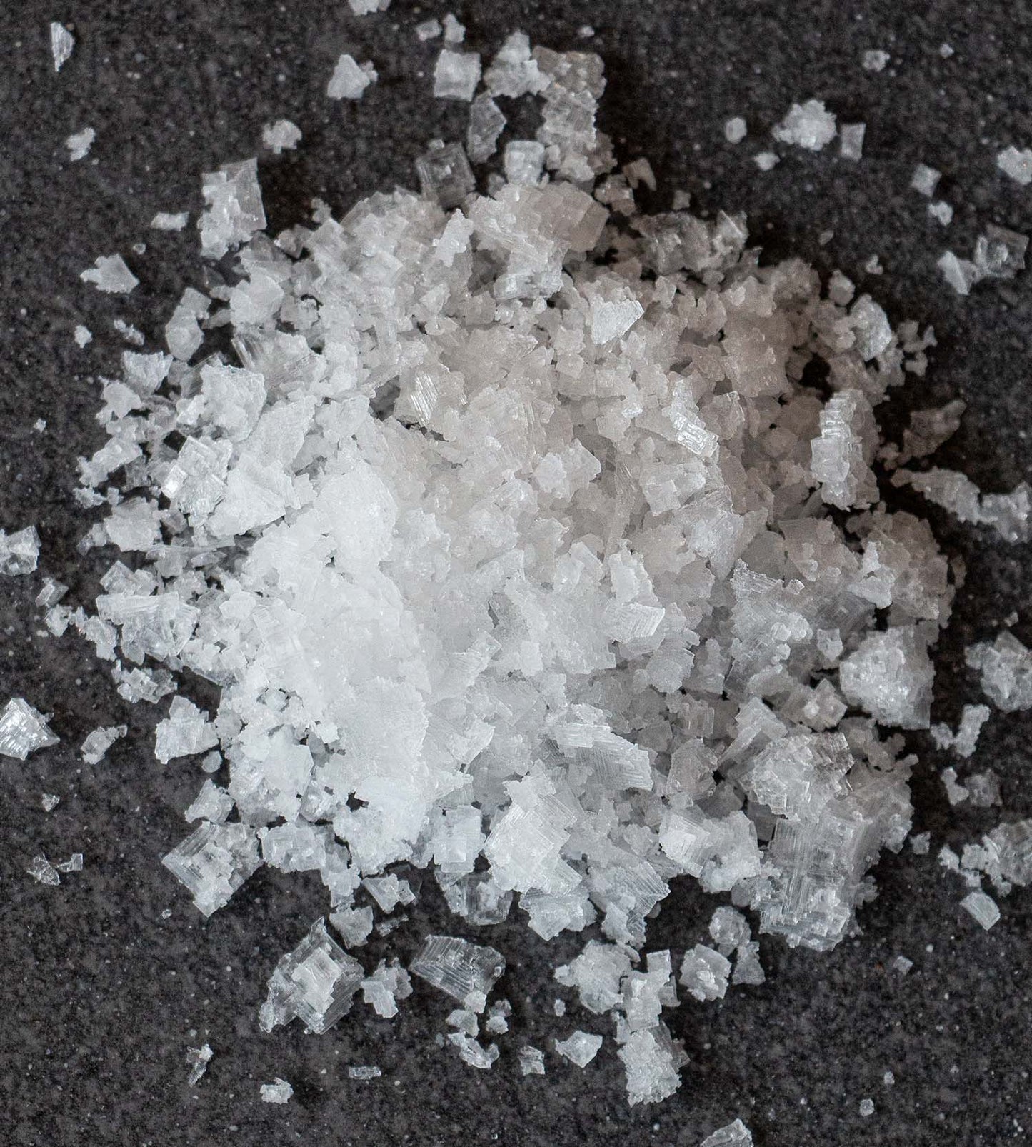 Closeup photo of a pile of white crystals of hand-harvested sea salt, on a dark grey tile background.