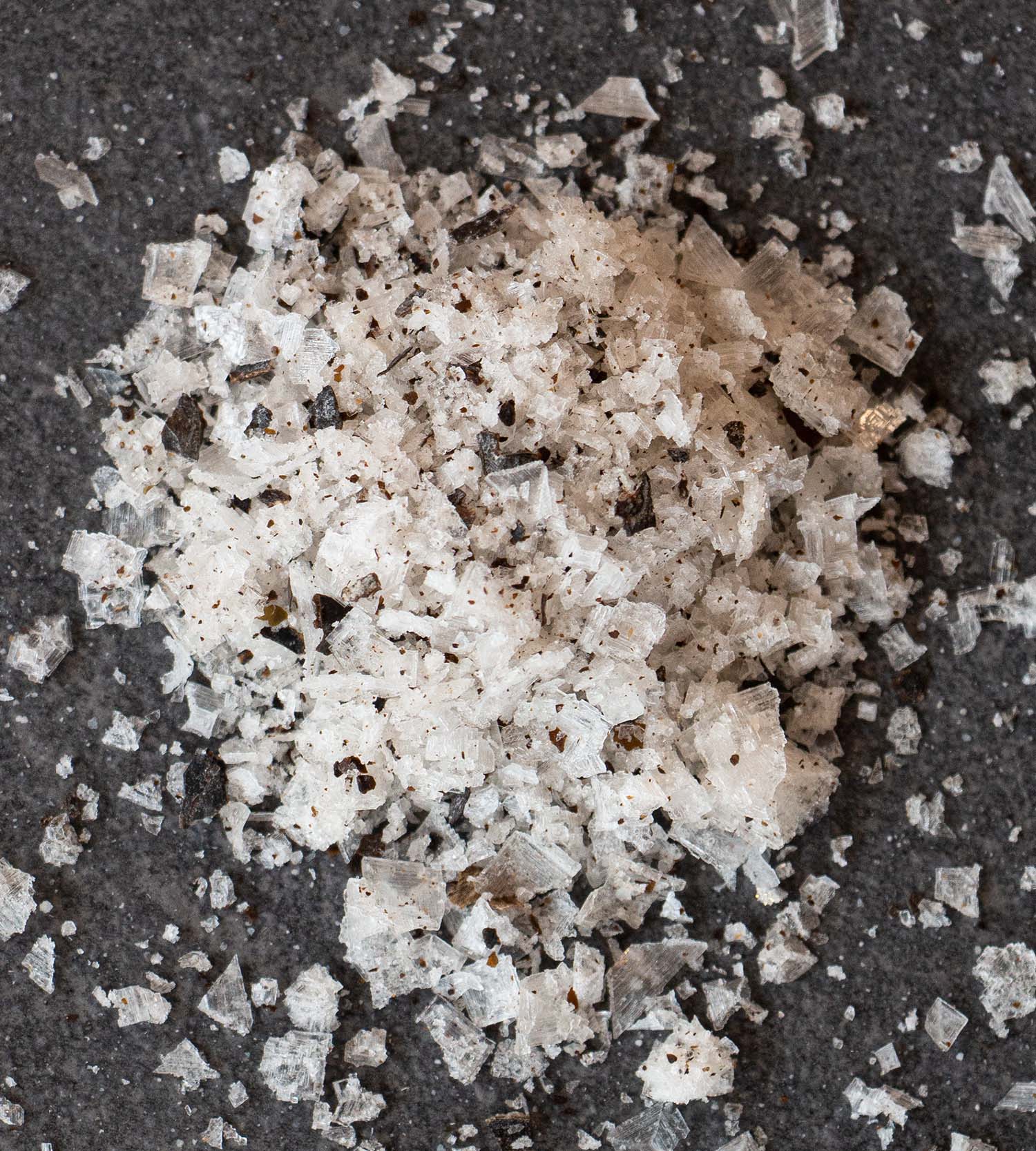 Closeup of a pile of white salt flakes interspersed with flakes of dried seaweed, against a dark grey slate background.