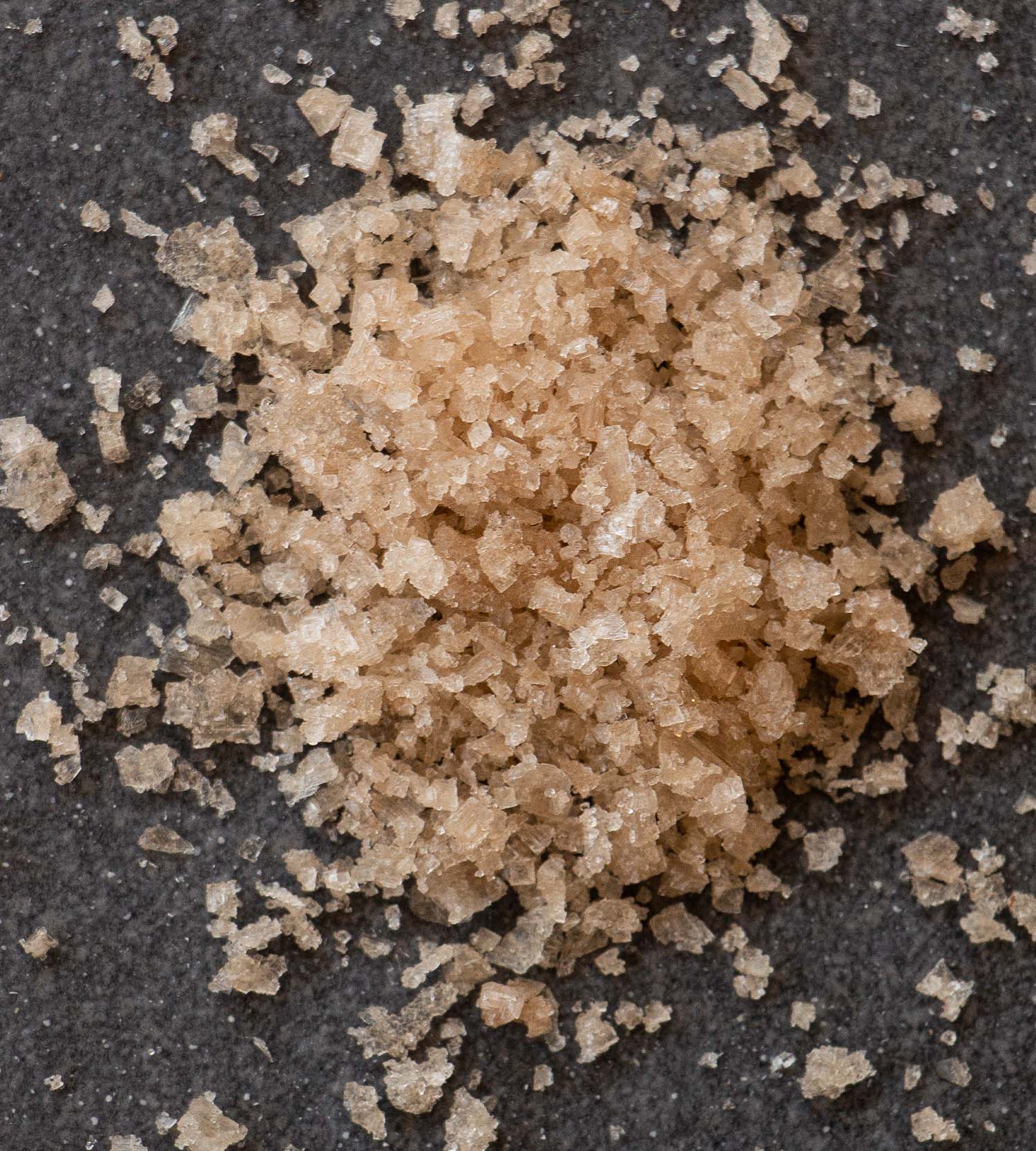 Closeup of a pile of light brown juniper-smoked sea salt flakes. The pile sits on a dark grey slate tile.